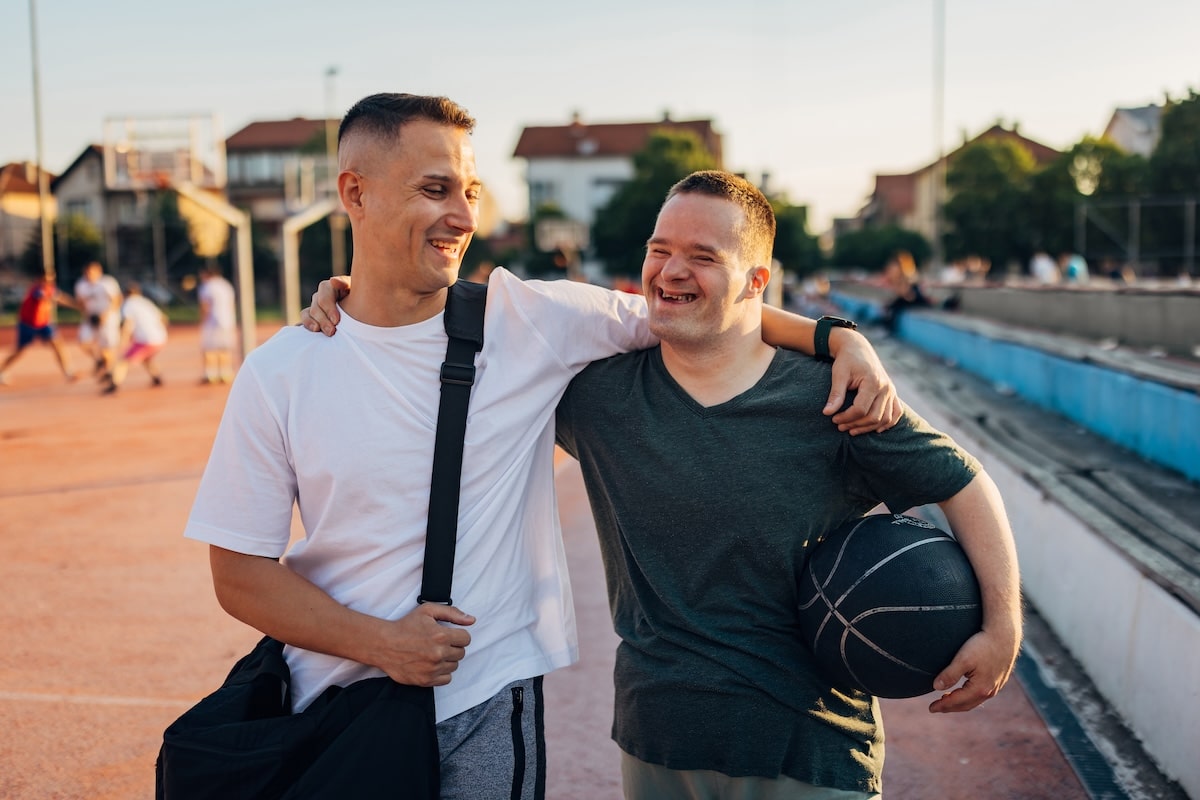 A man walking on a boardwalk with a disabled man, both smiling and with an arm around the other